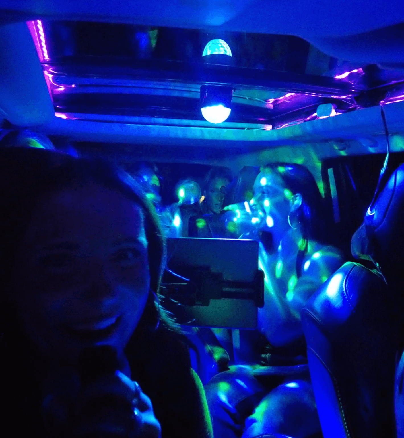 Women singing karaoke inside a car illuminated by vibrant blue and purple disco party lights.