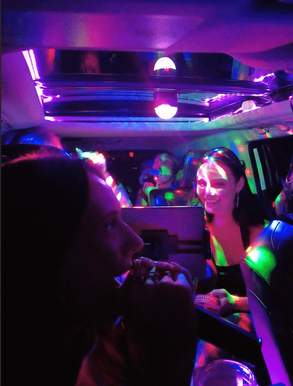 Women smiling inside a vehicle illuminated by vibrant, colorful disco party lights at night.