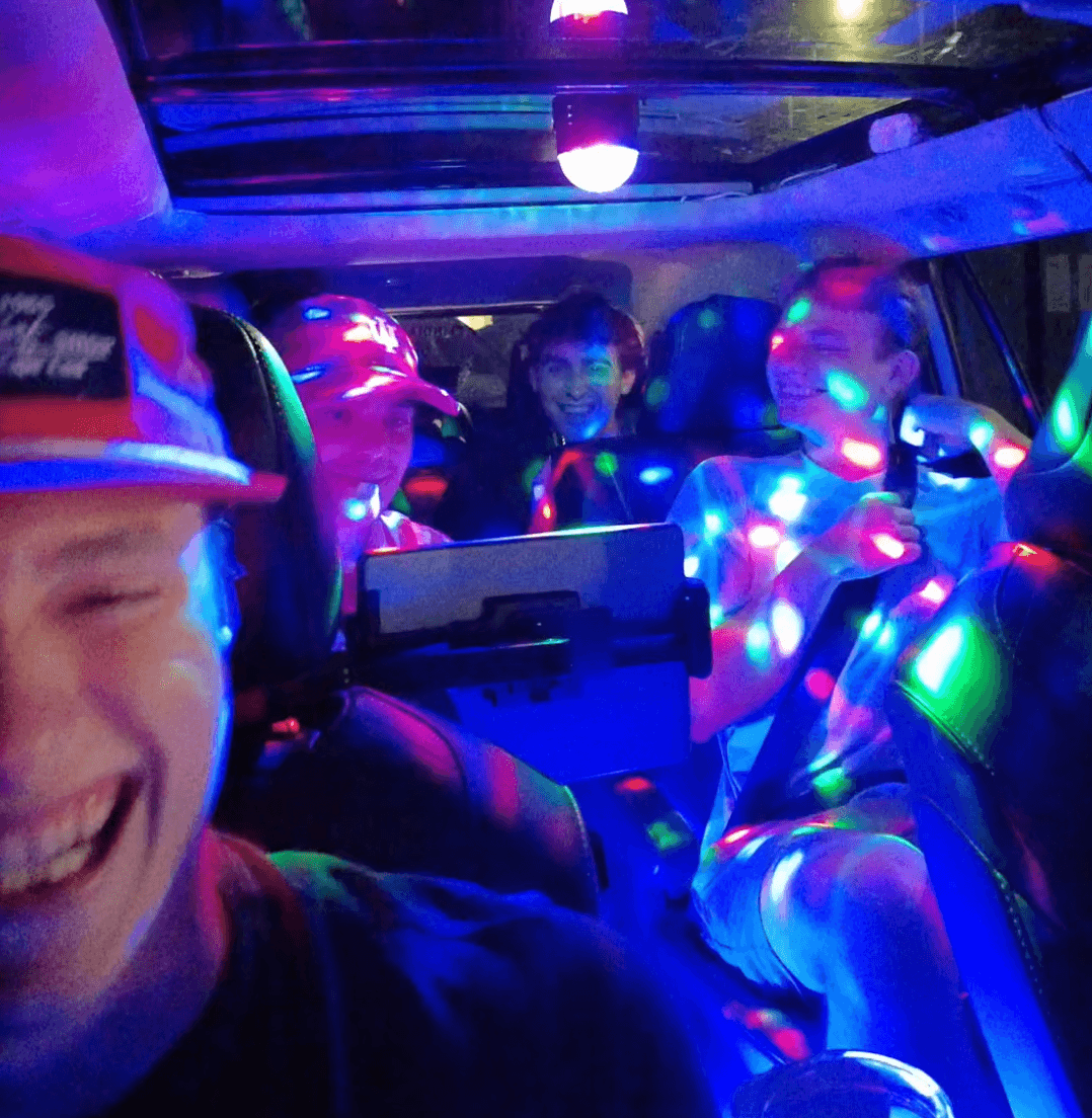 Four young men laughing inside a car illuminated by vibrant, colorful disco party lights.