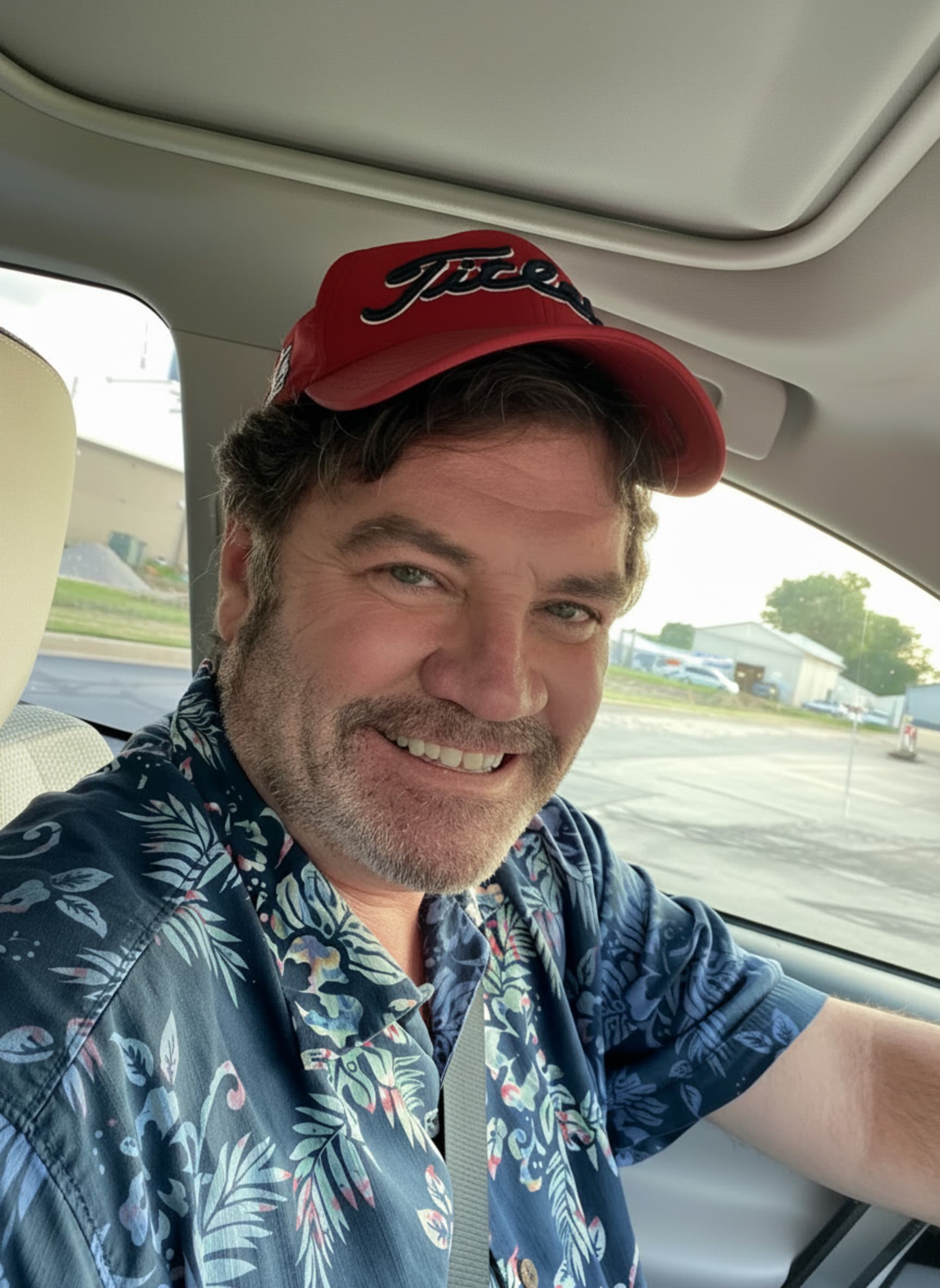 Smiling man with a beard, red Titleist cap, and blue floral shirt in a car.